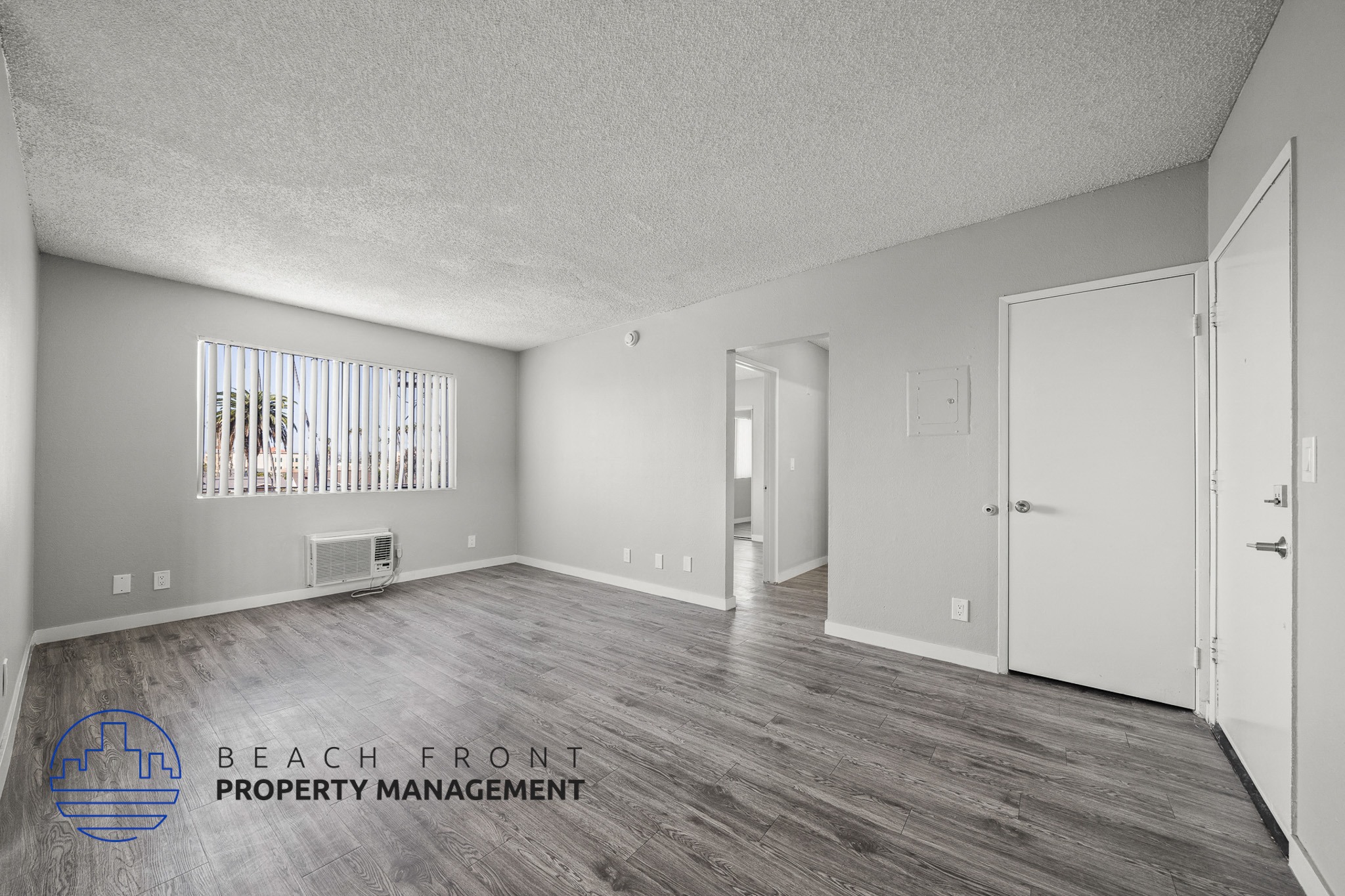A spacious room with wooden flooring and white walls, with a window covered by vertical blinds.